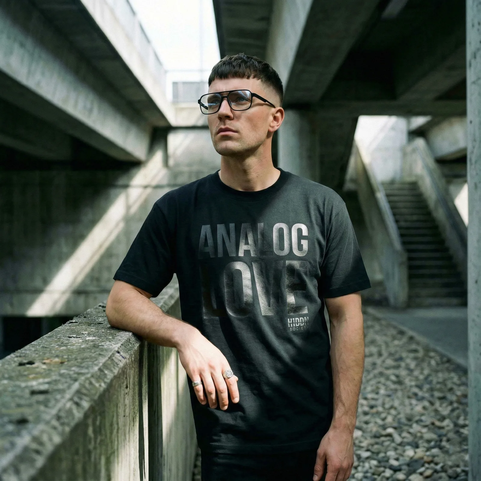 Person wearing a black H1DDN shirt beneath concrete structures